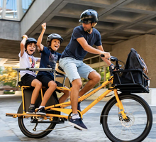 Load image into Gallery viewer, A photo of a Yuba Spicy Curry electric longtail cargo bike with the family pack installed, carrying two child passengers on the back of the bike.
