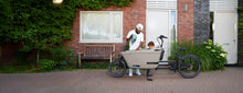 Load image into Gallery viewer, A lifestyle image of a Dolly Joy electric cargo bike parked in front of a house, with a child passenger sitting in the cargo box, while his father puts a helmet on the child's head.