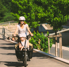 Load image into Gallery viewer, A lifestyle image of the Yuba Open Loader cargo box setup for the Yuba Supercargo CL electric cargo bike, shown in an urban setting. The bike is being ridden by a woman with a girl sitting in the front box.