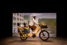 Load image into Gallery viewer, A studio image of a woman cycling a yellow Yuba Spicy Curry V4 electric longtail cargo bike with two child passengers sitting in the monkey bar add-on on the rear of the bike.