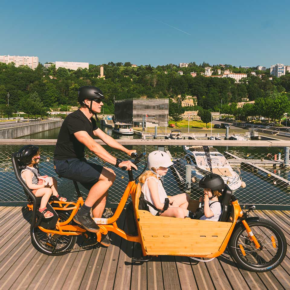 A lifestyle image showing the Yuba Supercargo CL electric cargo bike carrying three child passengers, with the 2be3 child seat inside the Bamboo Box and a baby seat on the rear carrier.