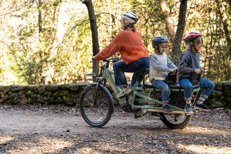 A lifestyle image showing a woman cycling a Yuba Spicy Curry+ cargo bike with two child passengers along a rural dirt track.