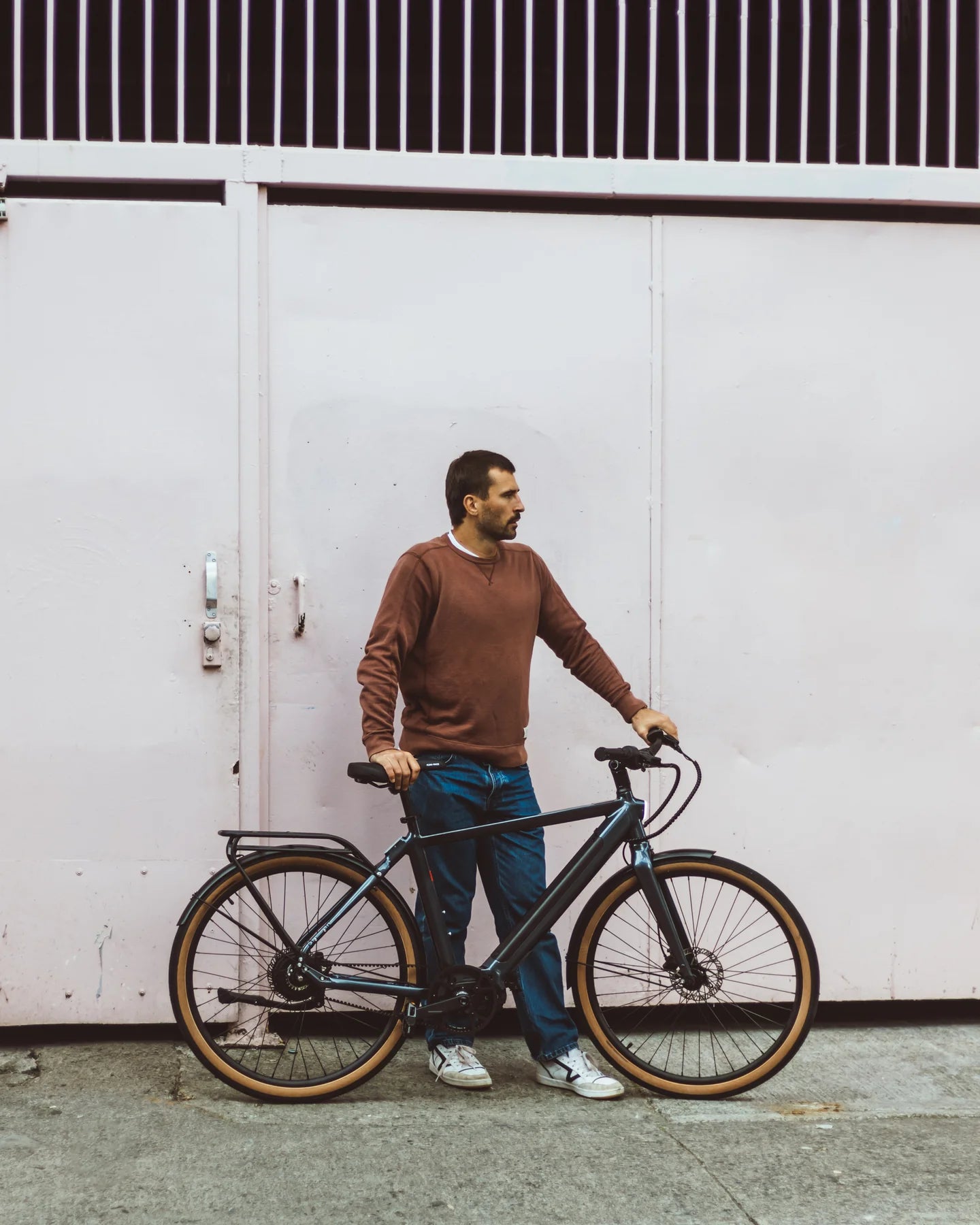 A lifetstyle image of the Kuma M1 electric bike with the bike parked against a white wall with a man standing beside the bike.