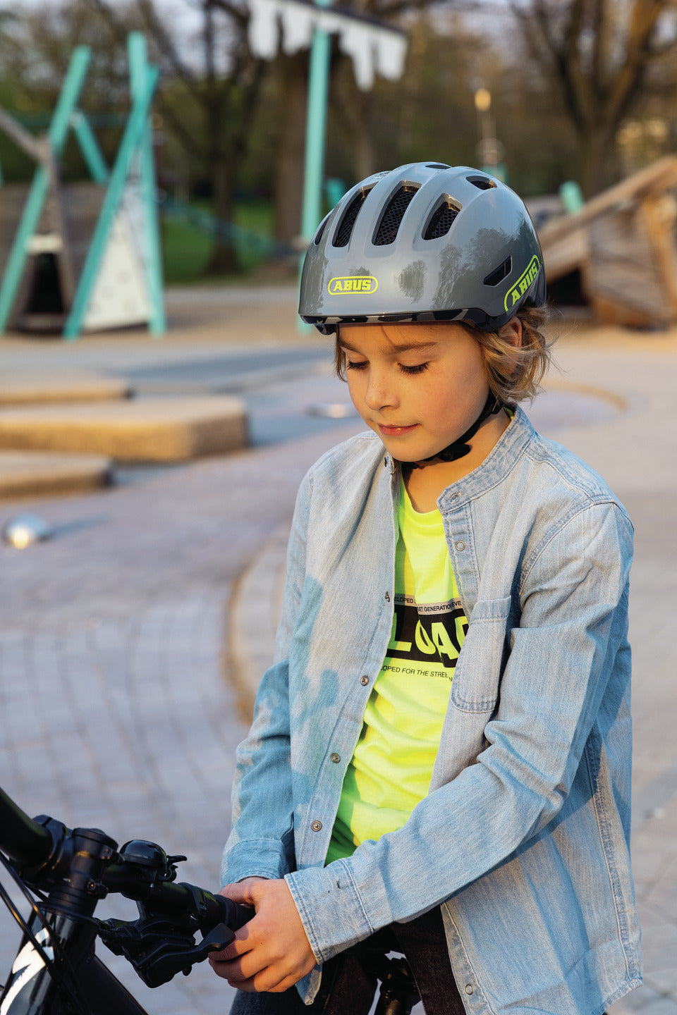 A lifestyle image of the Abus Smiley 3.0 kids cycle helmet being worn by a young boy cycling a bike in an urban playground.