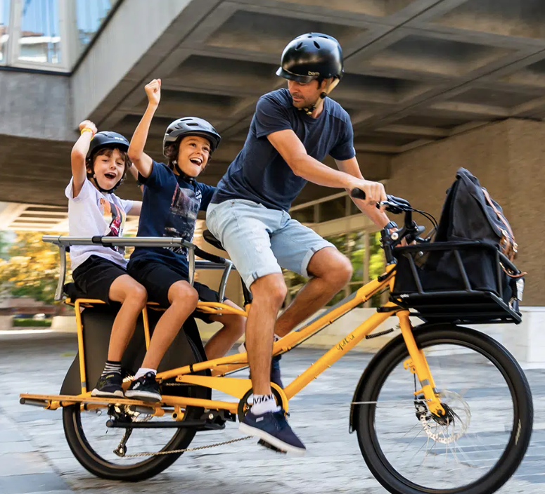 A photo of a Yuba Spicy Curry electric longtail cargo bike with the family pack installed, carrying two child passengers on the back of the bike.