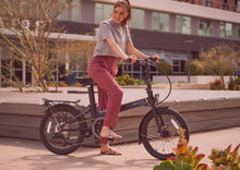 Load image into Gallery viewer, A lifestyle image of a Dahon Unio E20 folding electric bike with the frame colour obsidian. It is stopped in an urban space with a woman standing across it.

