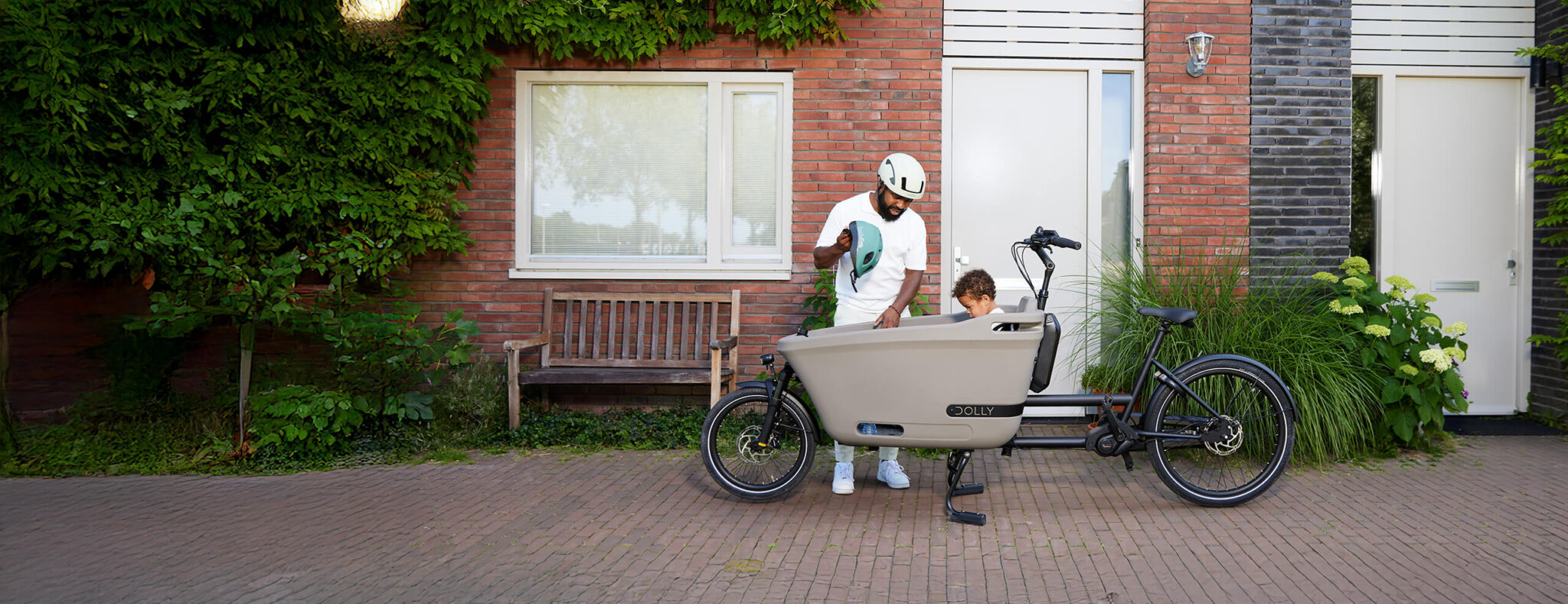 A lifestyle image of a Dolly Joy electric cargo bike parked in front of a house, with a child passenger sitting in the cargo box, while his father puts a helmet on the child's head. 