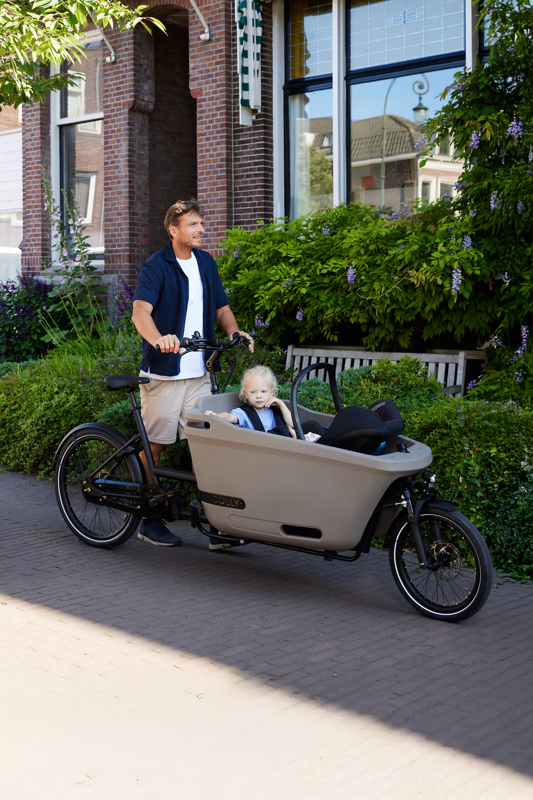 Lifestyle image of the Dolly Joy electric cargo bike showing a toddler and baby being transported in the cargo box. The parent is standing beside the cargo bike.