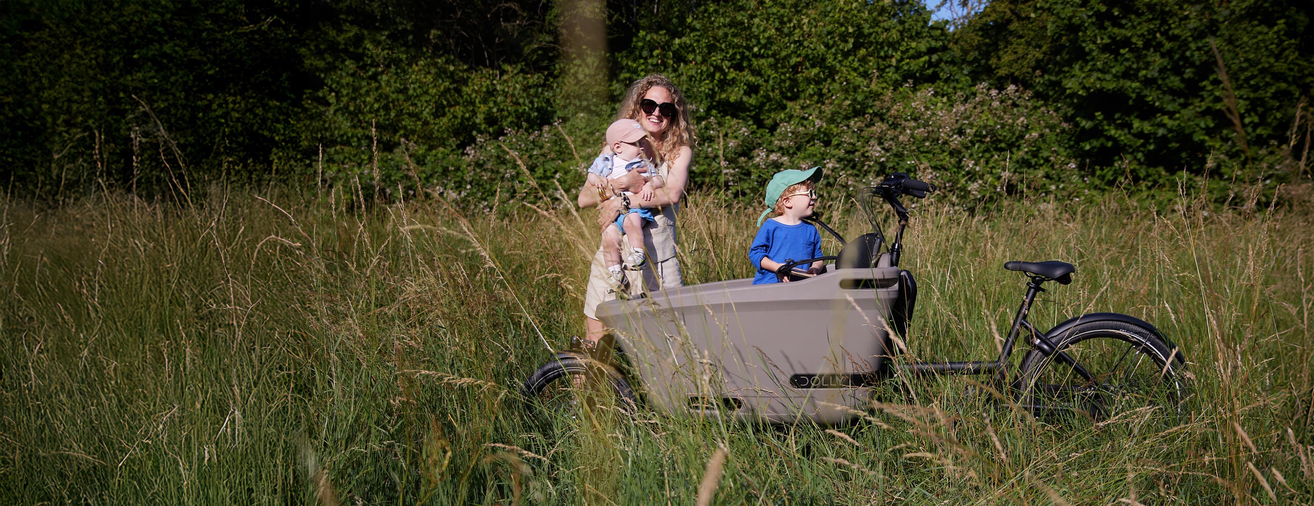 A lifestyle image of a Dolly Joy electric cargo bike parked in a grassy field, while a mother and her two children stand beside the bike. 