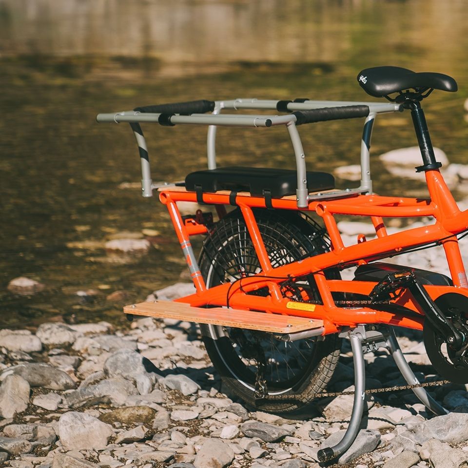 A photo of a Yuba Spicy Curry electric longtail cargo bike with the family pack installed. The bike is parked on a rocky shoreline beside a waterway. 