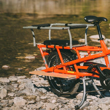 Load image into Gallery viewer, A photo of a Yuba Spicy Curry electric longtail cargo bike with the family pack installed. The bike is parked on a rocky shoreline beside a waterway. 
