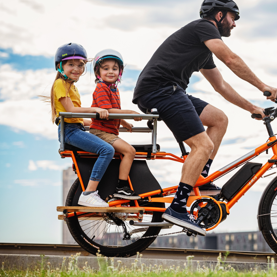 A photo of the Yuba Kombi E5 electric longtail cargo bike with two child passengers on the back. The bike is accessorised with the Yuba Kombi Family Pack.