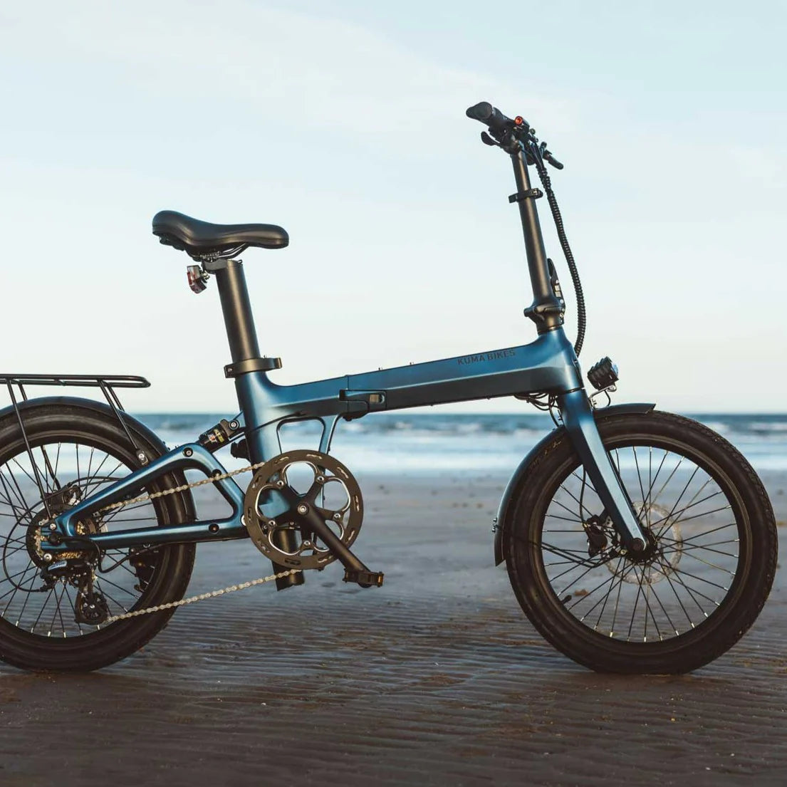 A photo of the Kuma F1 folding electric bike taken on a beach with the sea in the background. The light shows up the Matt Ocean Blue frame colour. The Kuma F1 is available to buy from Bleeper.