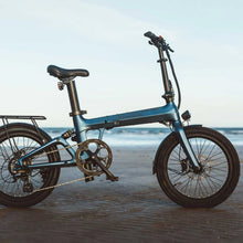Load image into Gallery viewer, A photo of the Kuma F1 folding electric bike taken on a beach with the sea in the background. The light shows up the Matt Ocean Blue frame colour. The Kuma F1 is available to buy from Bleeper.
