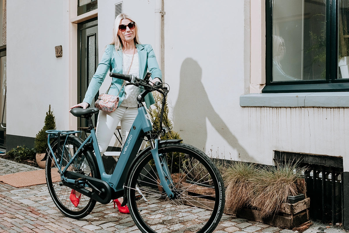A female model posing next to a Veloci Solid electric bike on an urban street. She has blonde hair and is wearing sunglasses and a green suit jacket, with bright red high heeled shoes. 