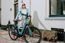 Load image into Gallery viewer, A female model posing next to a Veloci Solid electric bike on an urban street. She has blonde hair and is wearing sunglasses and a green suit jacket, with bright red high heeled shoes. 
