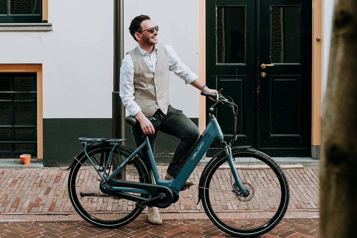 A male model posing next to a Veloci Solid electric bike on an urban street. He is wearing sunglasses, a beige waistcoat and slip-on shoes.