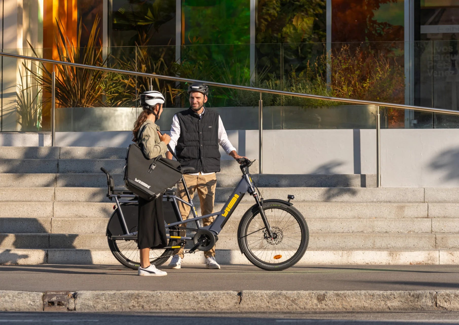 A lifestyle image showing a man and woman standing beside their Yuba Boda Boda longtail electric cargo bike. The woman is carrying a Yuba Eco Baguette bike bag using its shoulder strap.