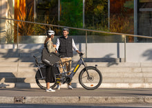 Load image into Gallery viewer, A lifestyle image showing a man and woman standing beside their Yuba Boda Boda longtail electric cargo bike. The woman is carrying a Yuba Eco Baguette bike bag using its shoulder strap.
