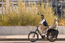 Load image into Gallery viewer, A lifestyle image of a man and a woman cycling a Yuba Boda Boda longtail electric cargo bike in an urban setting.
