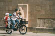 Load image into Gallery viewer, A lifestyle image showing a woman cycling a Yuba Kombi E6 electric longtail cargo bike through an urban setting. She is carrying two child passengers on the rear carrier rack of the bike. 
