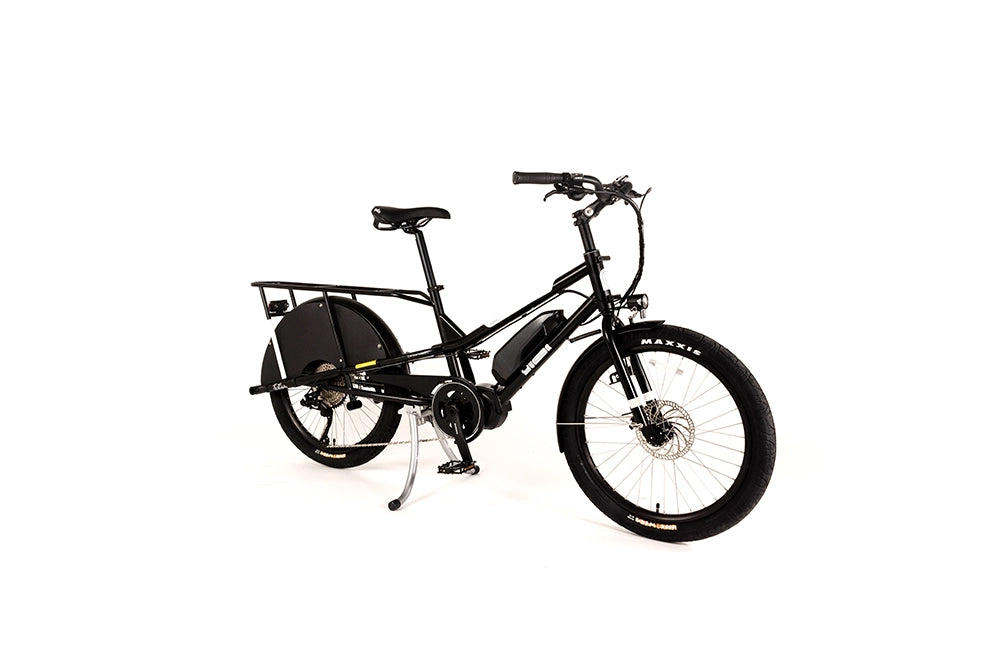 A product image of the Yuba Kombi E6 electric longtail cargo bike taken at an oblique angle from the front right of the bike. This is the bare layout of the bike, with no accessories added. The bike has a black frame. 
