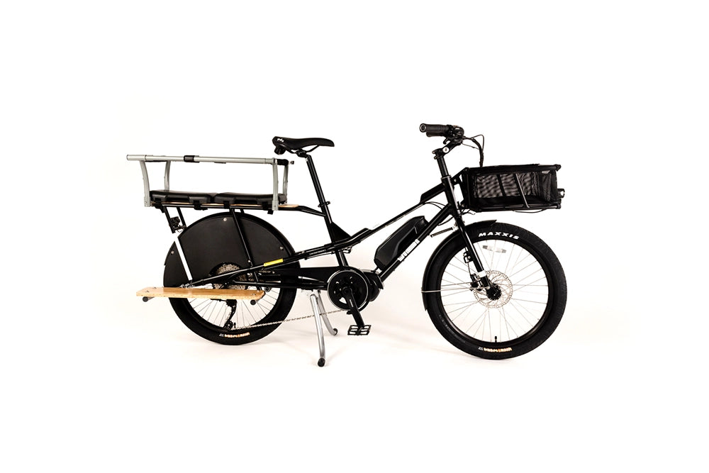 A product image of the Yuba Kombi E6 electric longtail cargo bike. The bike has a rear monkey bar, seat pads and footboard added, along with a basket on the front. The bike has a black frame. 