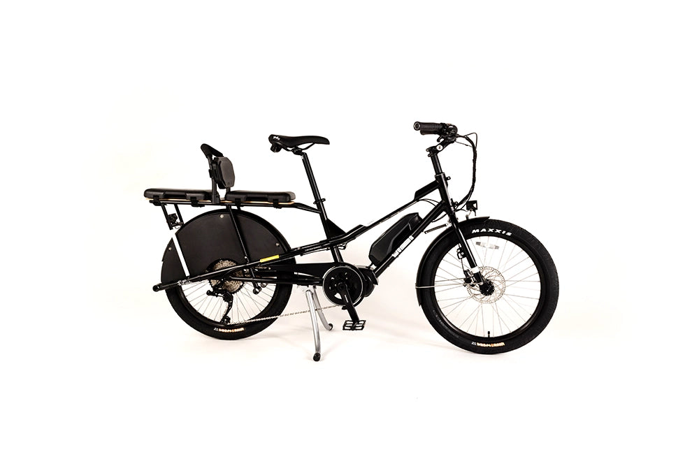 A product image of the Yuba Kombi E6 electric longtail cargo bike taken at an oblique angle from the front right of the bike. The bike has two seat pads and a back rest attached to the rear carrier rack. The bike has a black frame. 