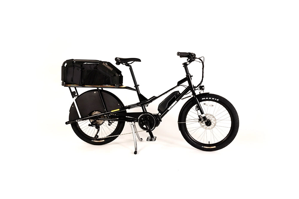 A product image of the Yuba Kombi E6 electric longtail cargo bike taken at an oblique angle from the front right of the bike. The bike has a wooftop basket attached to the rear rack, which is designed for carrying canine passengers. The bike has a black frame. 
