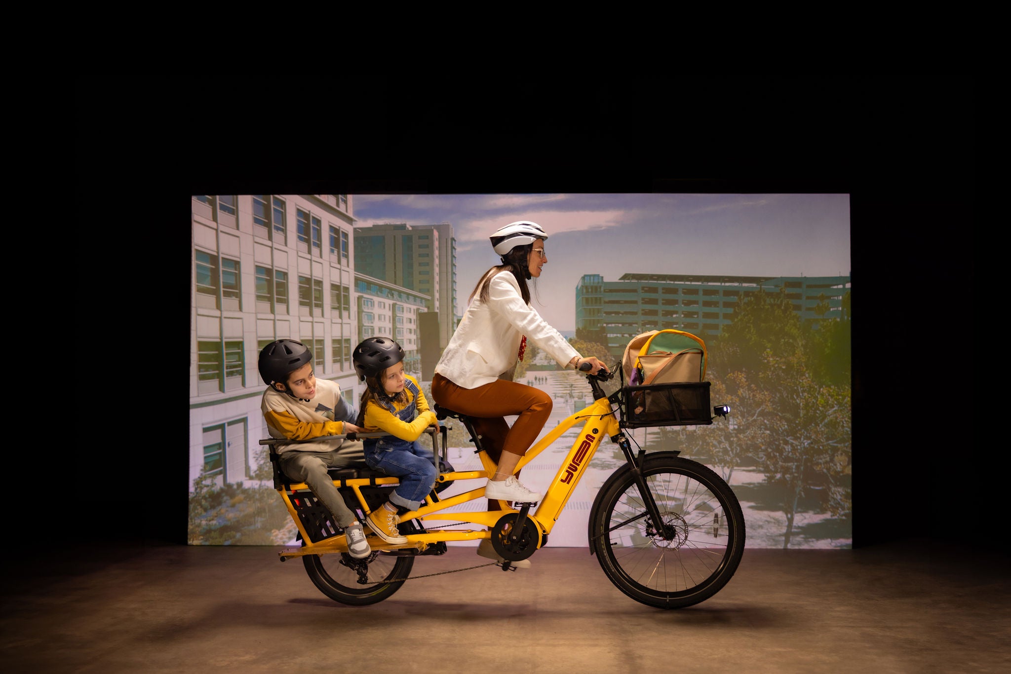 A studio image of a woman cycling a yellow Yuba Spicy Curry V4 electric longtail cargo bike with two child passengers sitting in the monkey bar add-on on the rear of the bike.