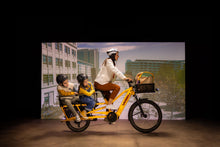 Load image into Gallery viewer, A studio image of a woman cycling a yellow Yuba Spicy Curry V4 electric longtail cargo bike with two child passengers sitting in the monkey bar add-on on the rear of the bike.
