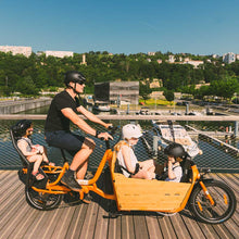 Load image into Gallery viewer, A lifestyle image showing the Yuba Supercargo CL electric cargo bike carrying three child passengers, with the 2be3 child seat inside the Bamboo Box and a baby seat on the rear carrier. 
