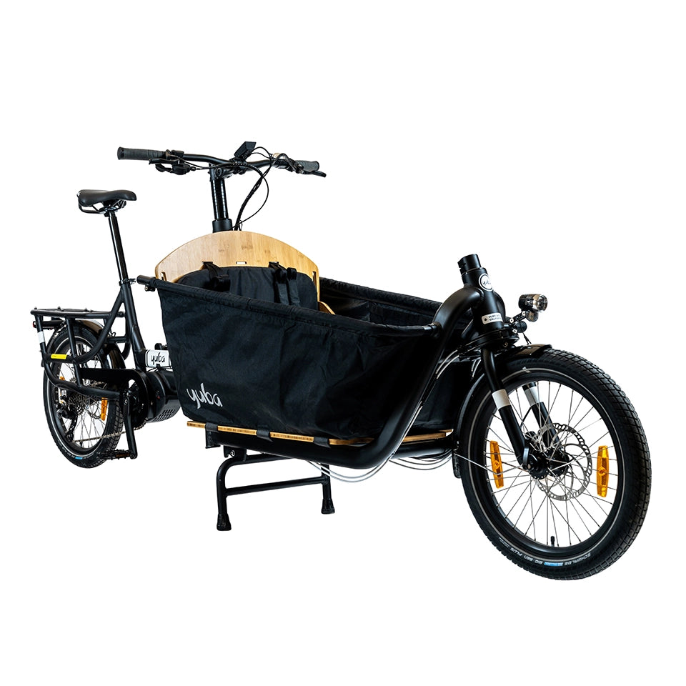 A product image of the Yuba Supercargo CL electric front-loading cargo bike. The model shown has a black frame, a canvas box with child seats at the front. The bike is available to buy from Bleeper.