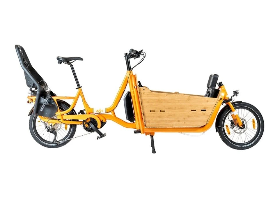 A product image of the Yuba Supercargo CL electric front-loading cargo bike. The model shown has an orange frame, a bamboo box with child seats, and a child seat on the rear carrier. The bike is available to buy from Bleeper. 