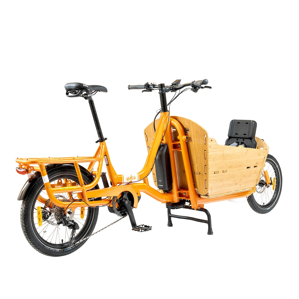 A product image of the Yuba Supercargo CL electric front-loading cargo bike. The model shown has an orange frame and a bamboo box with child seat at the front. The bike is available to buy from Bleeper.