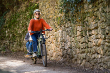 Load image into Gallery viewer, A lifestyle image showing a woman cycling a Yuba Spicy Curry+ cargo bike with two child passengers along a rural dirt track alongside an old stone wall.
