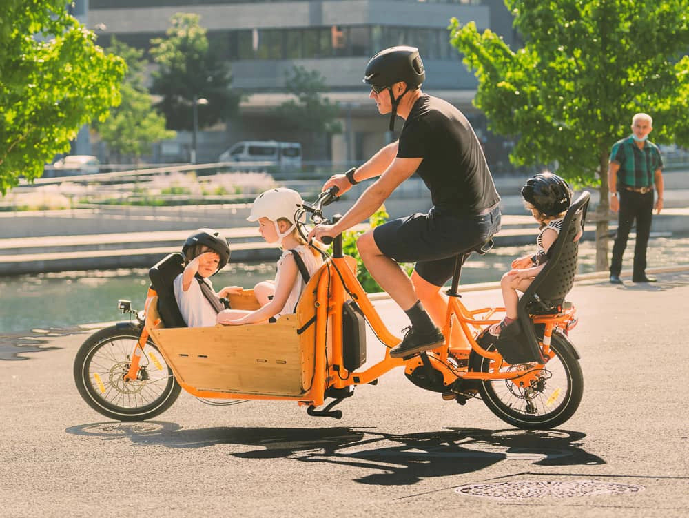 A lifestyle photo of the Yuba Supercargo CL electric cargo bike being cycled in an urban setting. A man is cycling the bike while two children sit in the front box and a third child is seated on the rear carrier of the bike. 