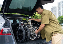 Load image into Gallery viewer, Man loading two folding Dahon bicycles into the boot of a car with a cityscape in the background
