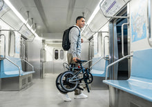 Load image into Gallery viewer, Man with a backpack and folding bike inside a subway train
