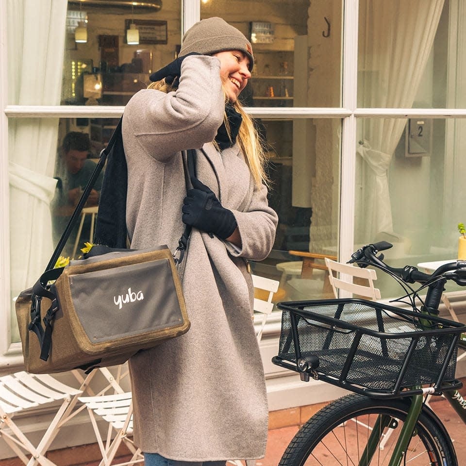 A photo of the Yuba Grab and Go bag being carried over the shoulder by a woman who is standing beside the front basket of a Yuba longtail cargo bike.