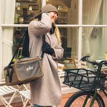 Load image into Gallery viewer, A photo of the Yuba Grab and Go bag being carried over the shoulder by a woman who is standing beside the front basket of a Yuba longtail cargo bike.
