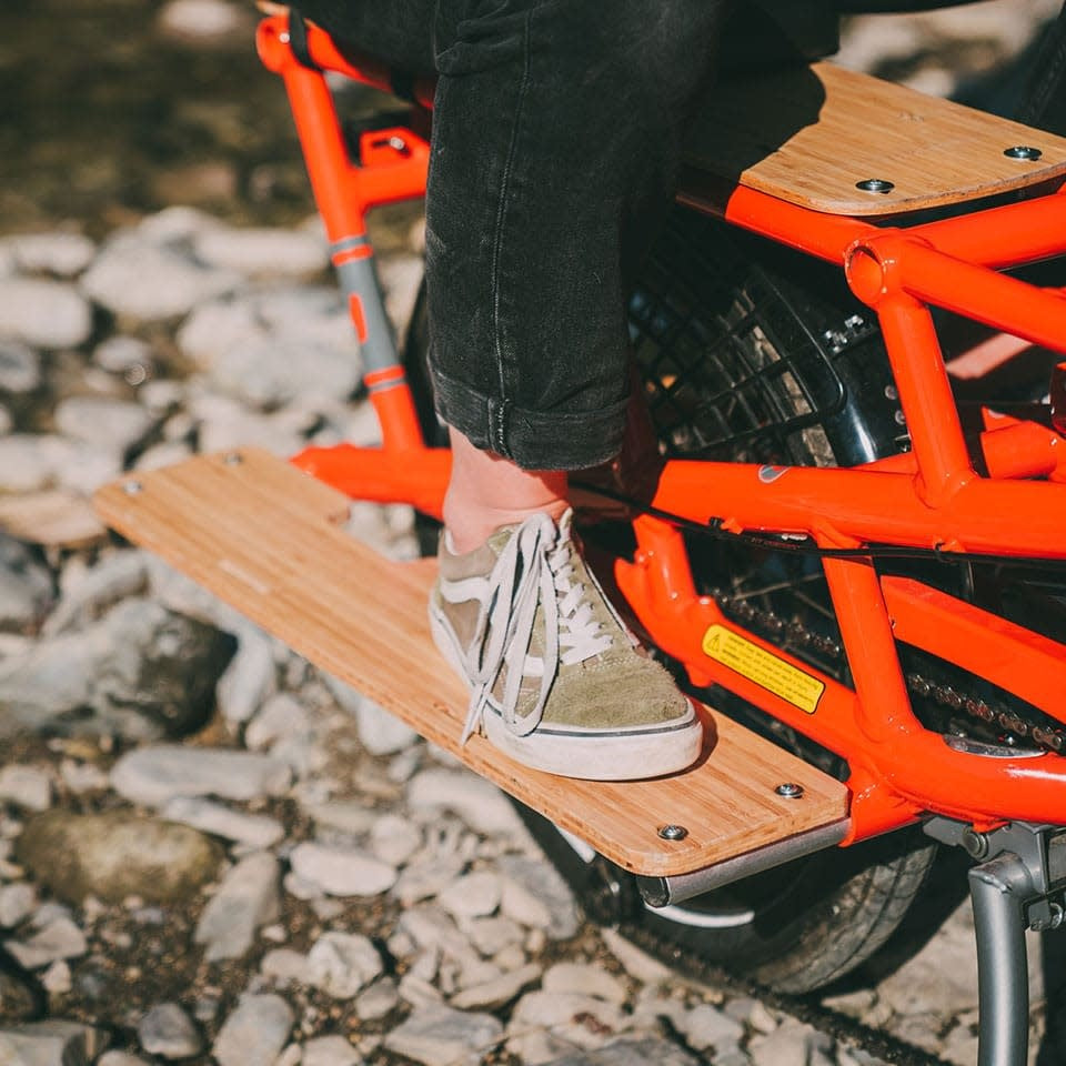 Product image showing a pair of Yuba Bamboo Sideboards for the Yuba Spicy Curry Electric Longtail Cargo Bike, with a rear passenger resting their feet on the sideboards.