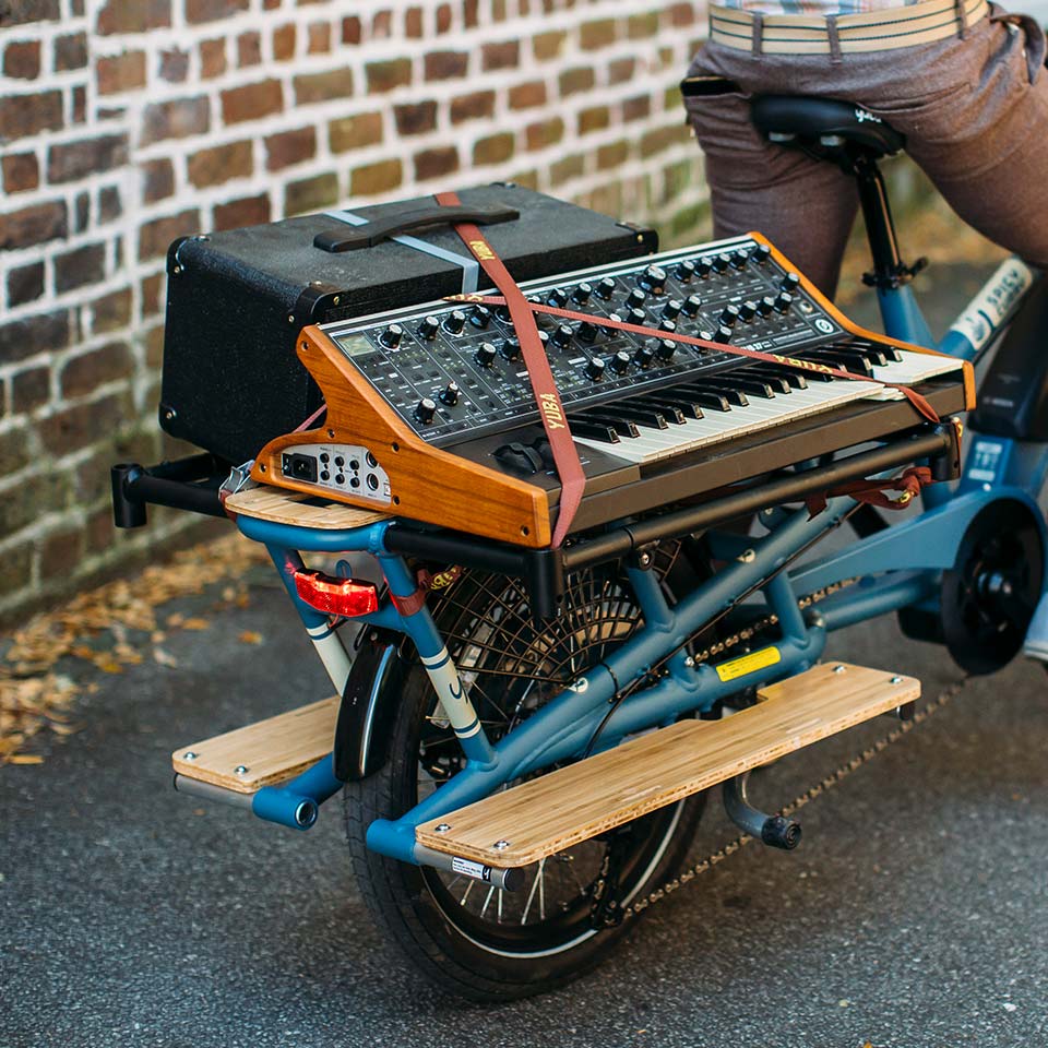 A product picture of the Yuba Carry-On platform which extends the carrying capacity of the Yuba Spicy Curry electric longtail cargo bike. A keyboard and a loudspeaker are strapped to the Carry-On rack.