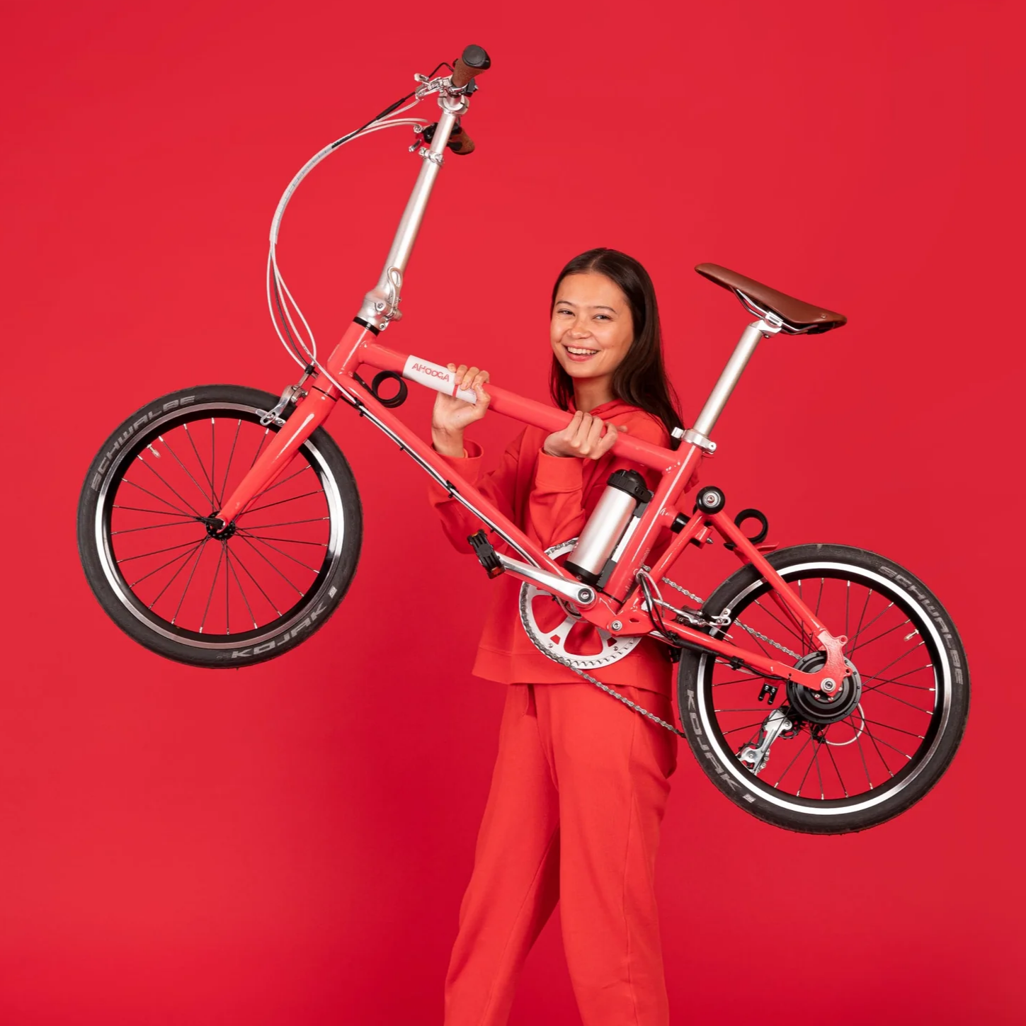 A product image of the Ahooga Active 24V folding electric bike being held up by a female model in a red studio setting. The frame colour is red. The Ahooga Active is available to buy from Bleeper.